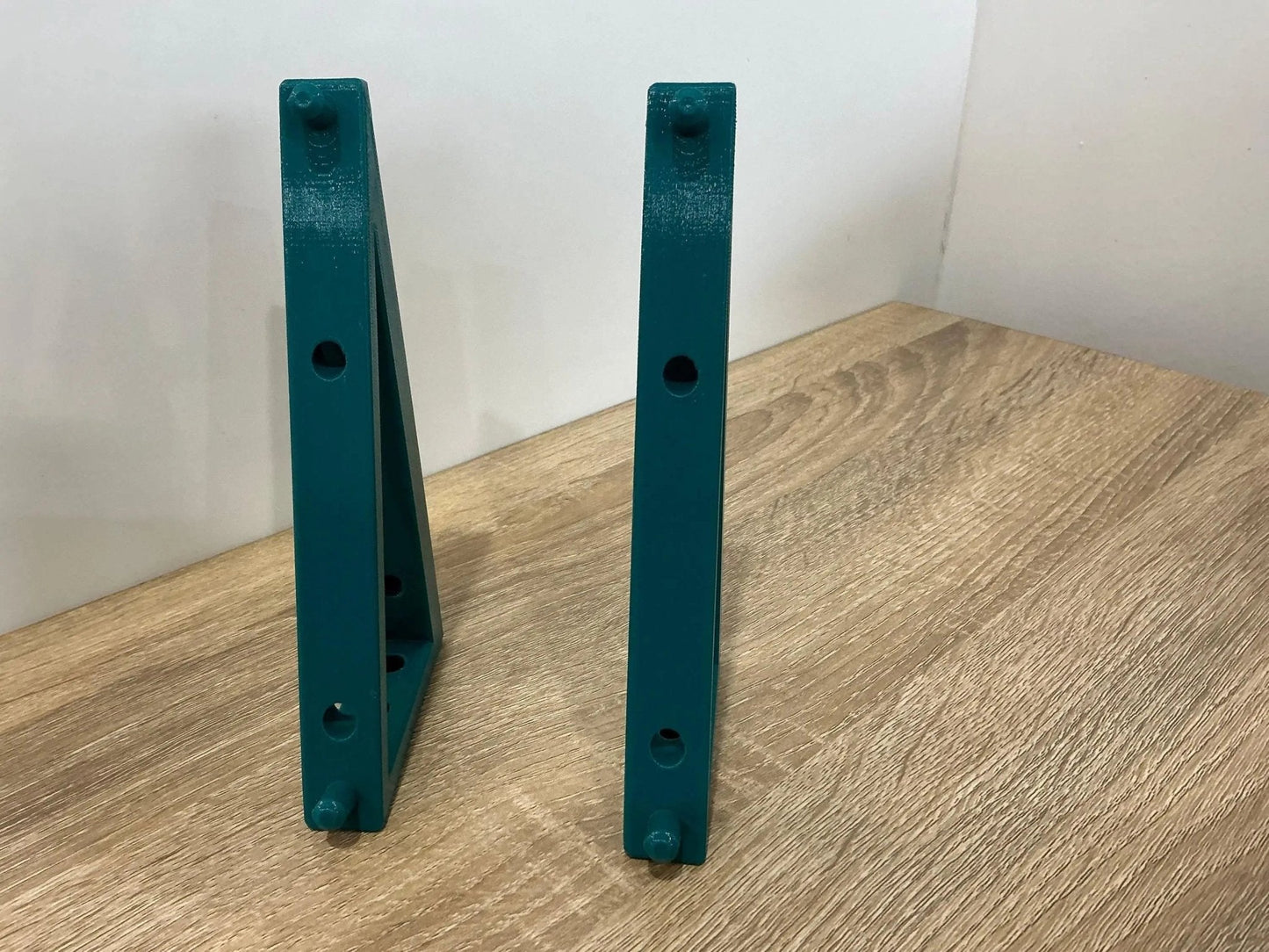 Teal-colored Makita 18V Dual Charger Wall Mount brackets positioned side by side on a wooden tabletop, illustrating their compact design.